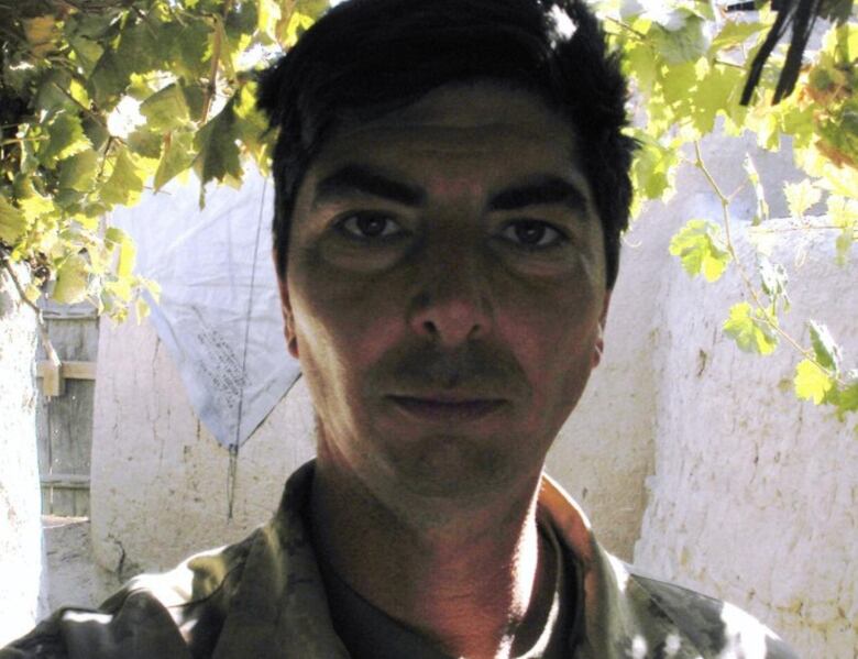 A young man in a military uniform gazes at the camera under a shelter of leaves. 