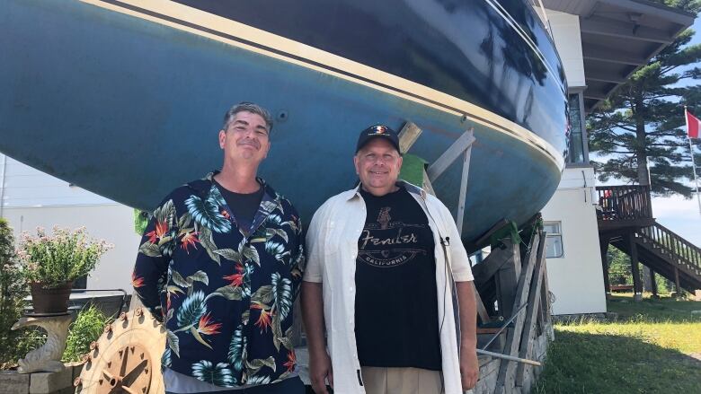 Two men stand smiling in front of a dry-docked boat. 