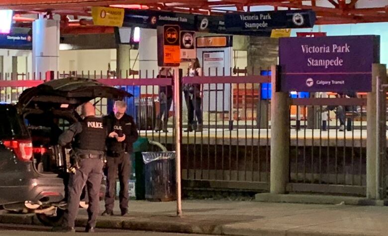  Two Calgary Police Service officers stand beside open hatch of vehicle on Macleod Trail while a pair of people wait for CTrain at Victoria Park Stampede station.