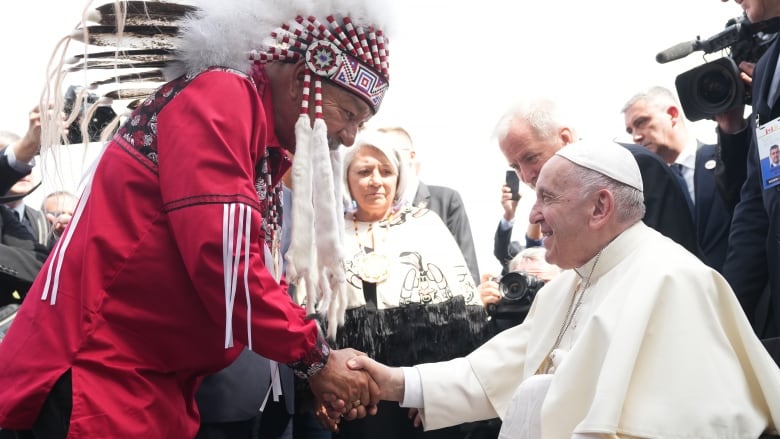 A man in a red shirt and traditional First Nations headdress, at left, greets Pope Francis, who is sitting in a wheelchair and dressed in white robes.