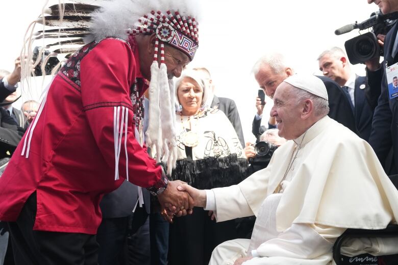 A man in a red shirt and traditional First Nations headdress, at left, greets Pope Francis, who is sitting in a wheelchair and dressed in white robes.