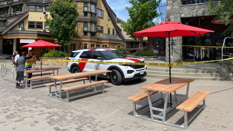 Yellow police tape encircle a white vehicle, which is parked in front of picnic tables and storefronts.