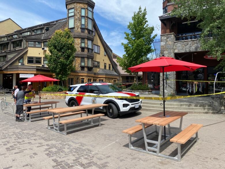 Yellow police tape encircle a white vehicle, which is parked in front of picnic tables and storefronts.