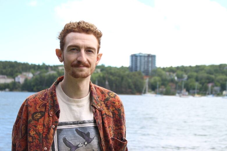 A person stands next to the water with trees, boats and buildings in the background.