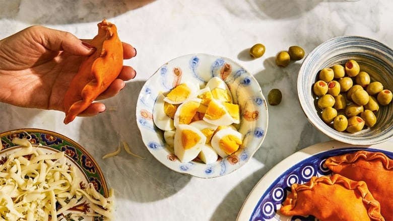 a hand holding an empanada that has just been stuffed. bowls of cheese, boiled eggs and olives sit in the background. 