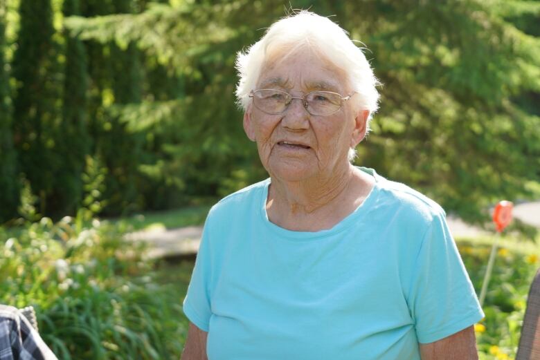 An elderly Mohawk woman with short white hair stands outside in the sun. 