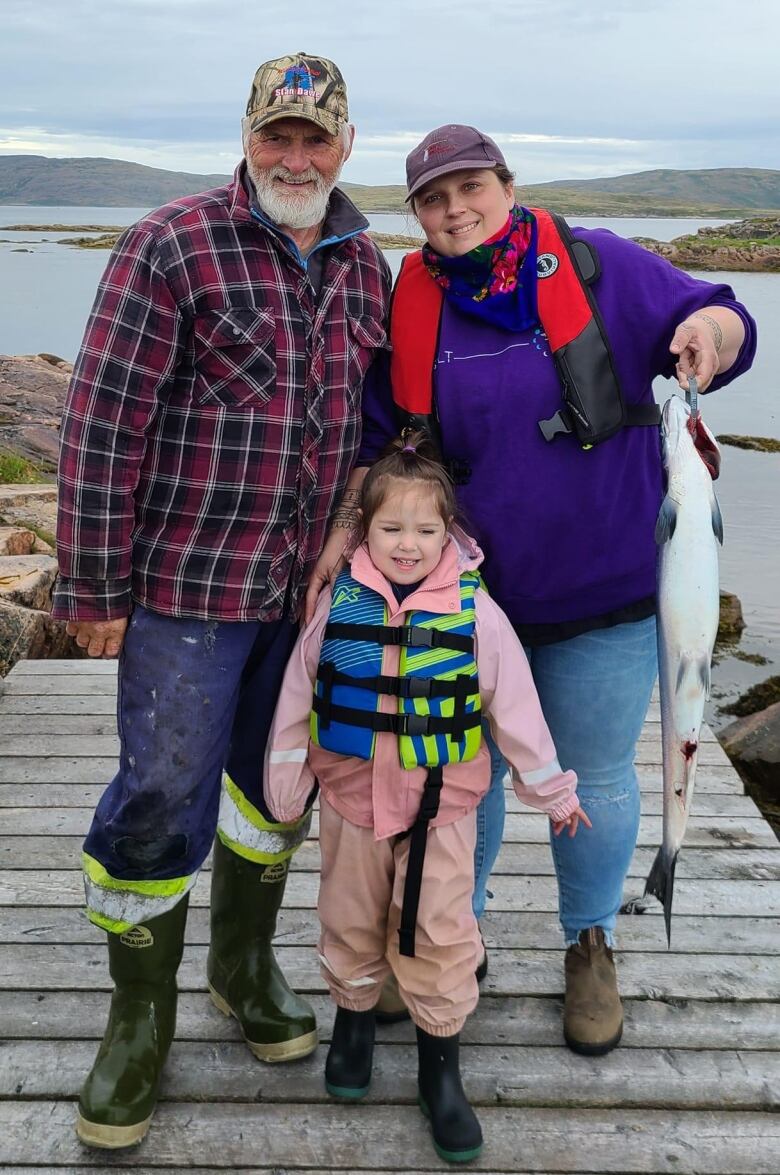 An older man, a girl and a woman stand on a boardwalk, with a lake behind them. The woman is holding a salmon.