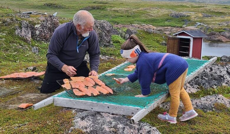 A young girls holds a piece of fresh salmon while an older man prepares a tray of salmon filets for smoking.