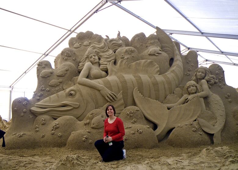 A woman in a red shirt is kneeling in front of a sand sculpture the size of a city bus, which features a mermaid, a massive fish, child mermaids, and is surrounded by a school of fish. 