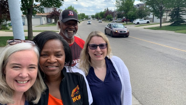 Four people stand together on a sidewalk along a busy residential road. 