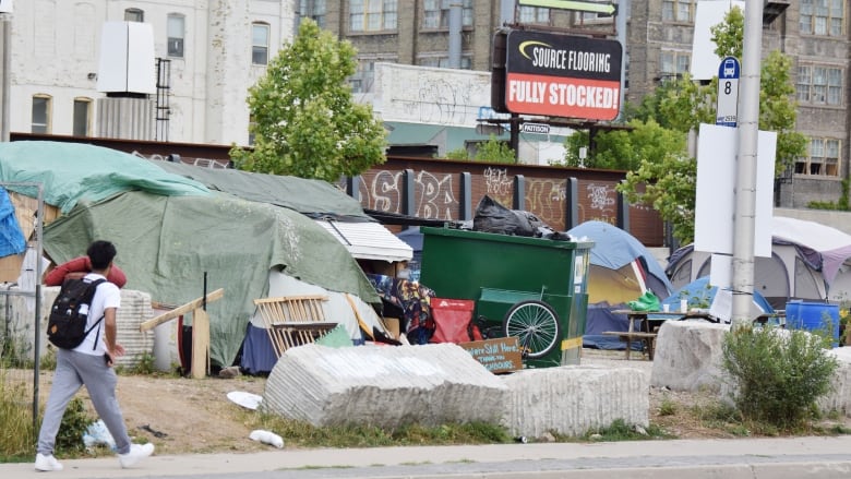 Man in white T-shirt, grey sweatpants, white running shoes and carrying a black backpack walks by vacant lot where people have set up tents. There's garbage, a folding chair and a dumpster visible.