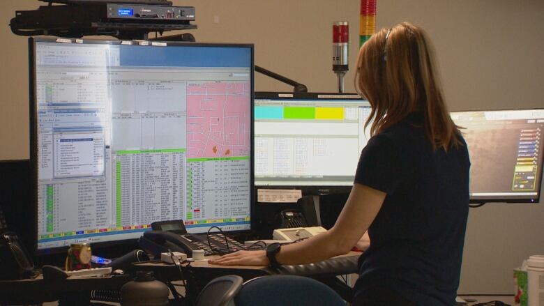 A woman with her back to the camera deals with a phone switchboard and multiple computer screens.