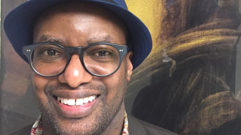 A Black man wearing glasses and a fedora smiling at the camera and standing in front of an abstract painting