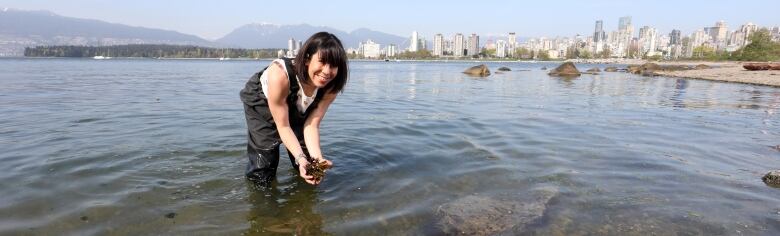 Angela, a white woman with long black hair, stands in thigh-high water and holds up some dirt.