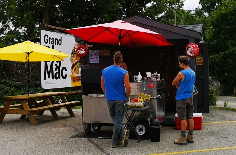 Two men stand in front of a hot dog cart. The silver cart has a red umbrella above it and is located in a parking lot with trees in the background.