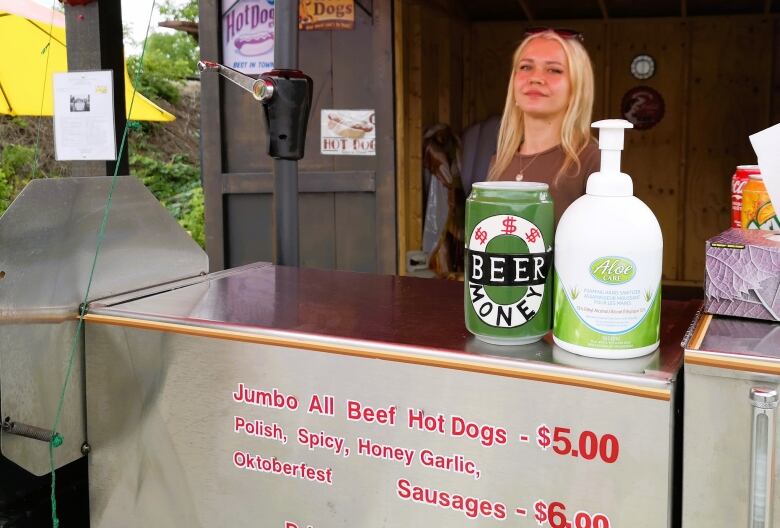 Young woman stands behind the counter of a hot dog cart where a sign advertises 'jumbo all beef hot dogs $5 or Oktoberfest sausages $6.