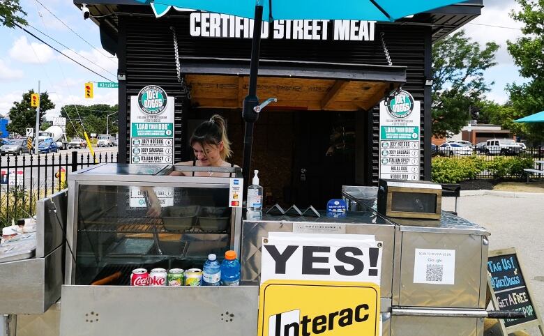 A young woman stands behind a hot dog cart with a bright teal umbrella and a sign in front says 