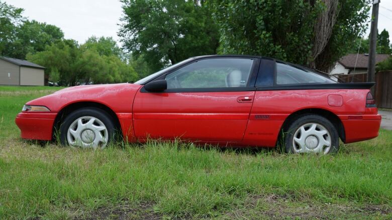 A red, two-door car with a black roof is parked on a grassy lot. The tires are deflated and the wheels sunk into the ground.