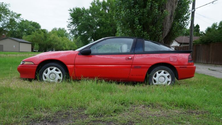 A red, two-door car with a black roof is parked on a grassy lot. The tires are deflated and the wheels sunk into the ground.