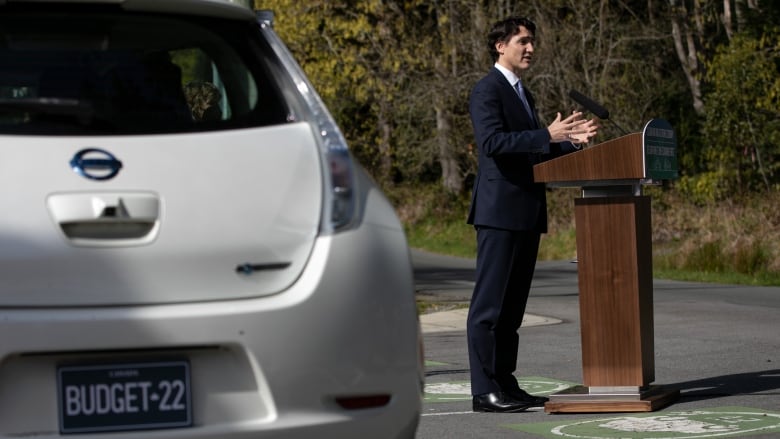 Prime Minister Justin Trudeau stands at a podium in a parking lot next to an electric car with 'Budget-22' written on the licence plate.