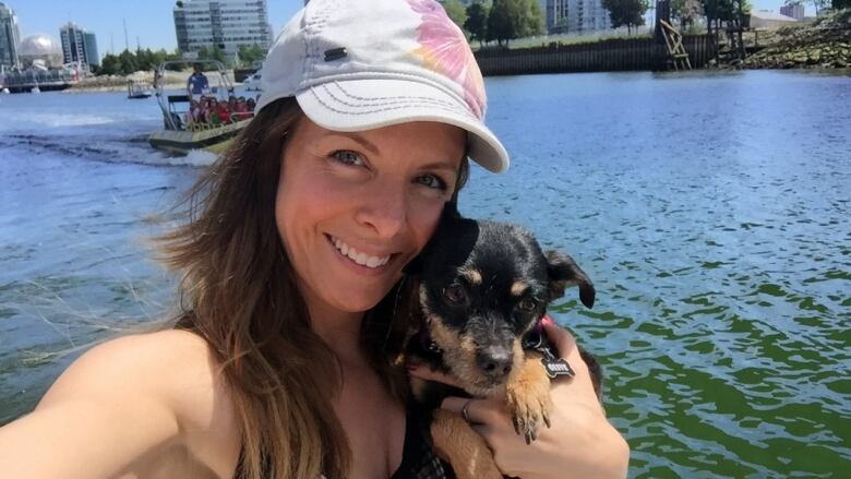 A selfie photo shows a woman with long brown hair under a white baseball cap holding a tiny black and brown dog. She is wearing a black tank top and positioned in front of False Creek in Vancouver, with the condo towers of Olympic Village in the background.