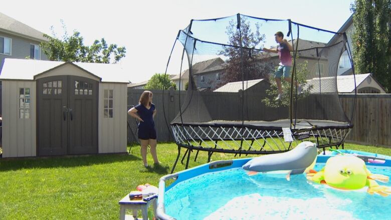 A mom watches her son jump on a trampoline in a backyard.