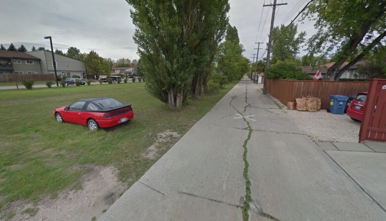 A red car sits in a grassy field next to a city back lane.