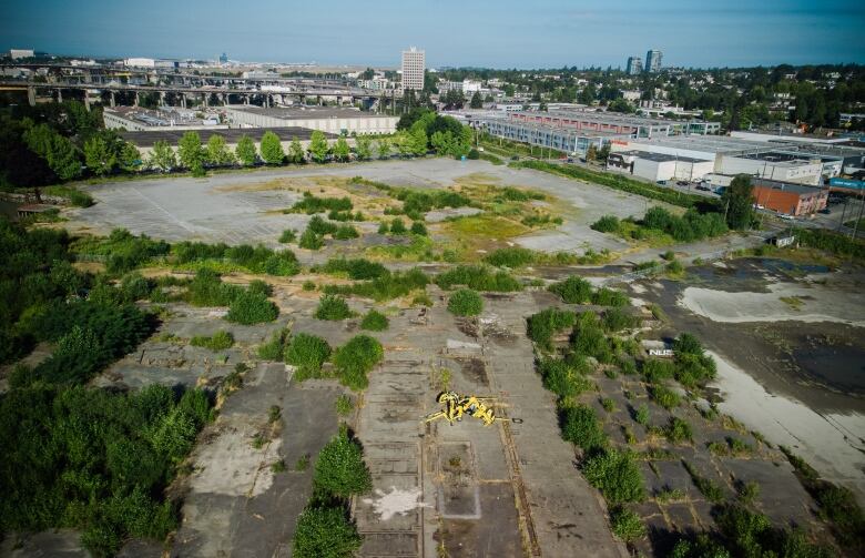 A drone picture showing a large yellow statue sitting in an empty land, with a few skyscrapers and industrial buildings visible in the background.