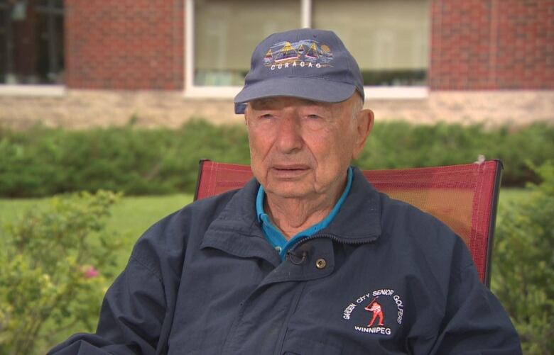 An elderly man wearing a ball cap and a jacket sits outsides on a chair.