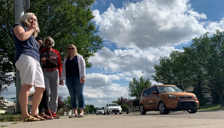 People stand beside a residential street with vehicles driving past.