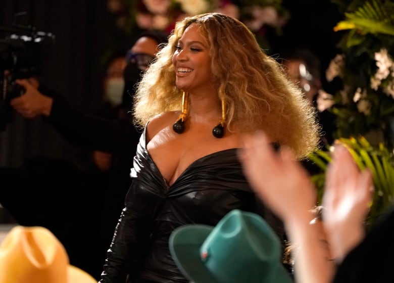 A buxom woman with a thick head of long, blond, braided hair, wearing a classy black dress, smiles widely at an applauding crowd.