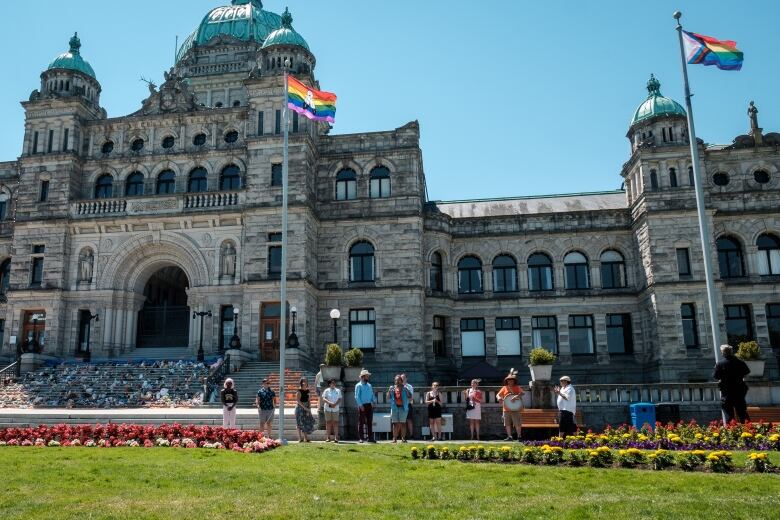 A group of people stand underneath the large B.C. Legislature building, with two rainbow Pride flags flying above them. One of the flags has two feathers on it, to commemorate two-spirit people.