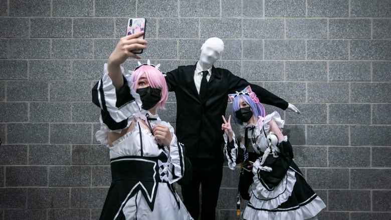 Two people in Japanese cosplay, wearing masks, take a selfie with a mannequin dressed in a suit.