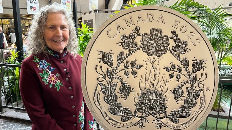 A woman in a beaded jacket stands next to a large silver coin that has a beadwork design on it. The coin says 