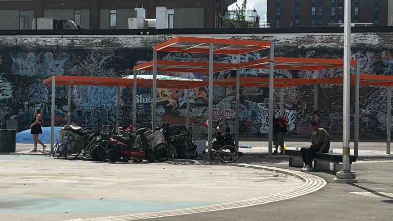 An orange shade overhang provides shade to a collection of bicycles. Behind, there is a wall covered in artistic grafitti. 