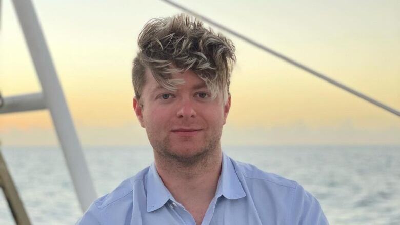 A man with swept-up yellow hair wearing a blue dress shirt gazes at the camera while the ocean rolls behind him. 