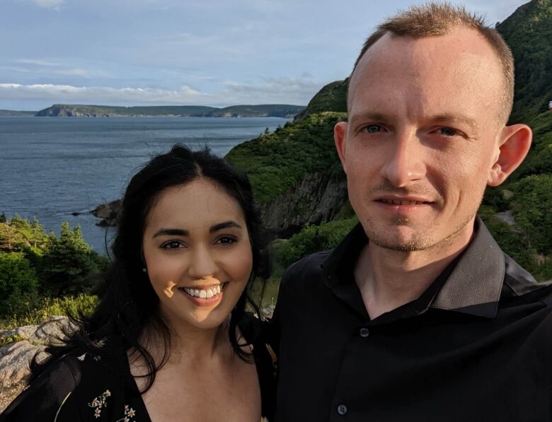 A woman and a man stand in the sun on a hill with the ocean behind them.