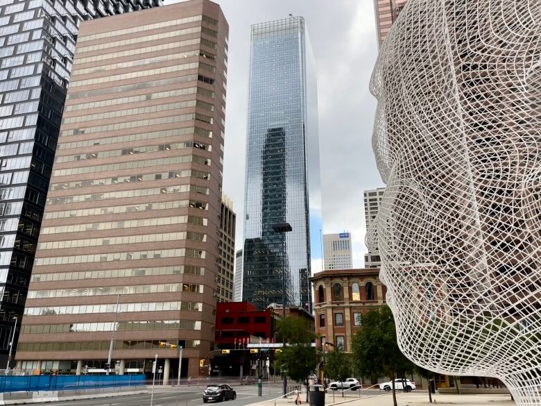 Tall buildings and downtown street. In the foreground is a white wire sculpture of a man's face.