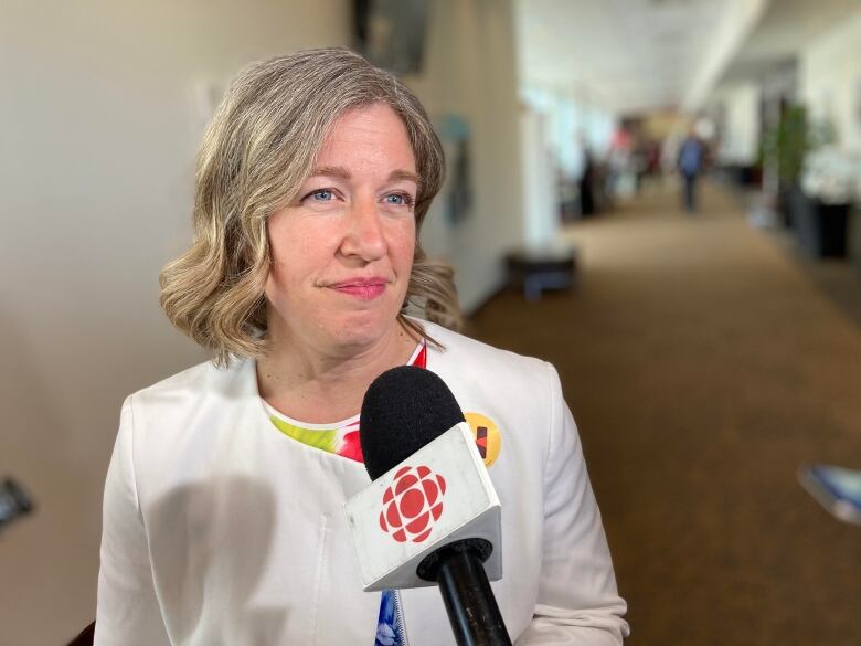 A woman with short blond hair and a white shirt standing in front of a CBC-branded microphone that is pointed at her