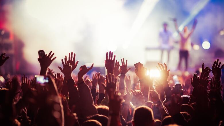 Concertgoers are seen from the back, raising their hands against bright concert stage spotlights. 