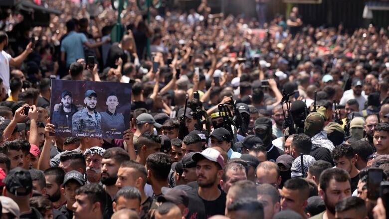 Swarms of people are shown outside, one holds up a placard with the pictures of three men.