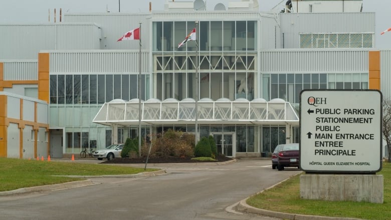 The Queen Elizabeth Hospital in Charlottetown, a white building with many windows. A sign out front points visitors to parking and the main entrance.