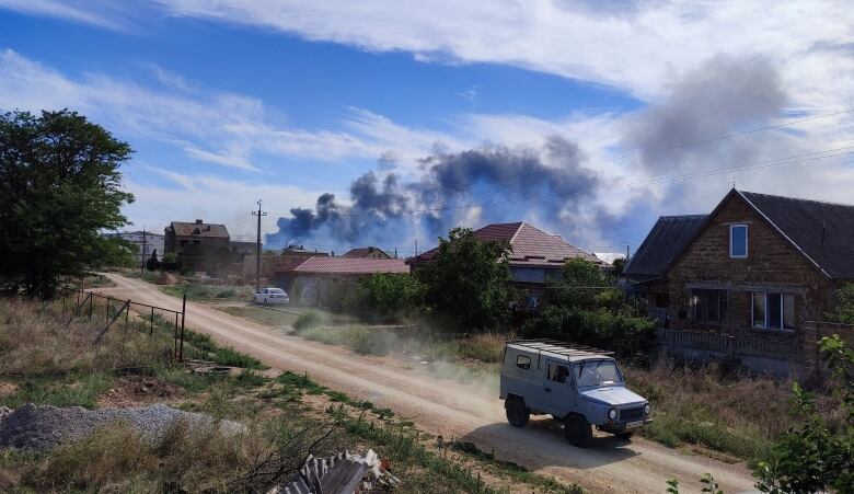 A vehicle driving past houses on a dirt road. Smoke is seen rising in the distance. 