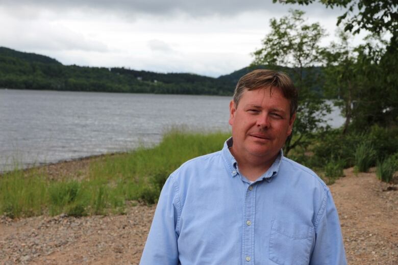 A middle-aged man in a blue collared shirt looks at the camera. He is standing on a rocky beach. 