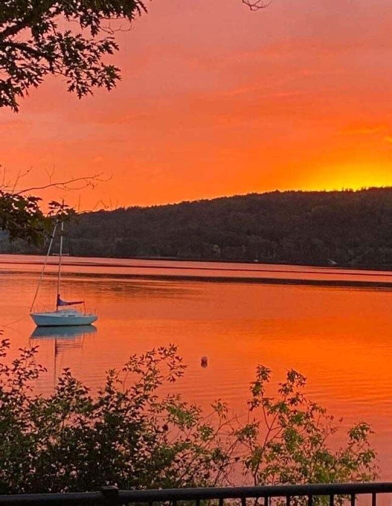 A setting sun over water lights the sky up with colours of orange and yellow. A sailboat is seen in the distance. 