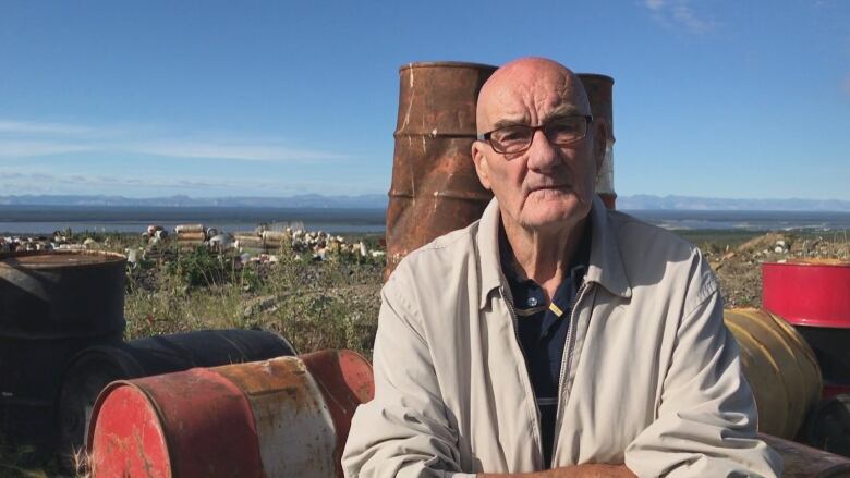 A man sits outside by some old oil drums.