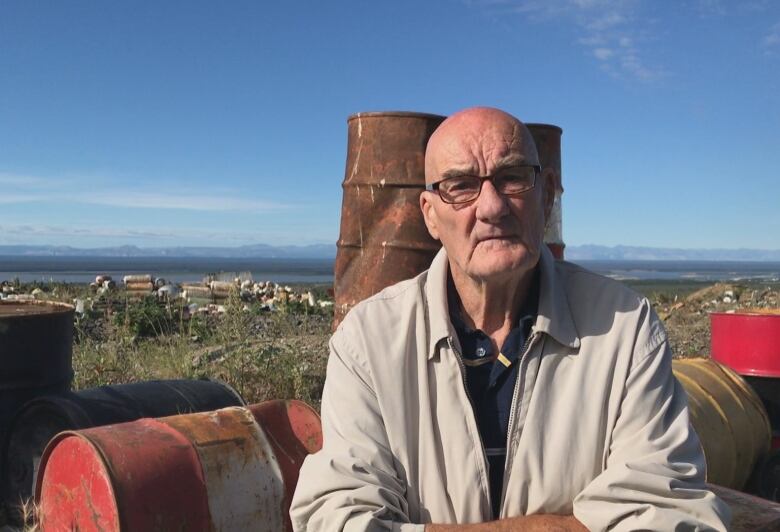 A man sits outside by some old oil drums.