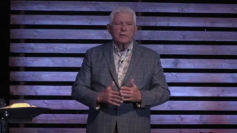 A man with grey hair wearing a suit stands on a stage.