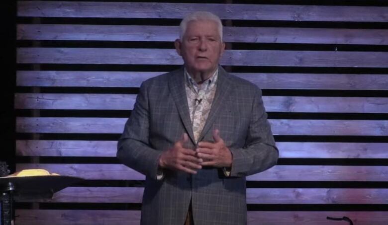 A man with grey hair wearing a suit stands on a stage.