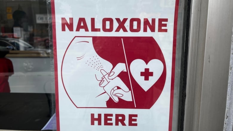 A sign in a window says Naloxone here and has an illustration of a nasal spray. 